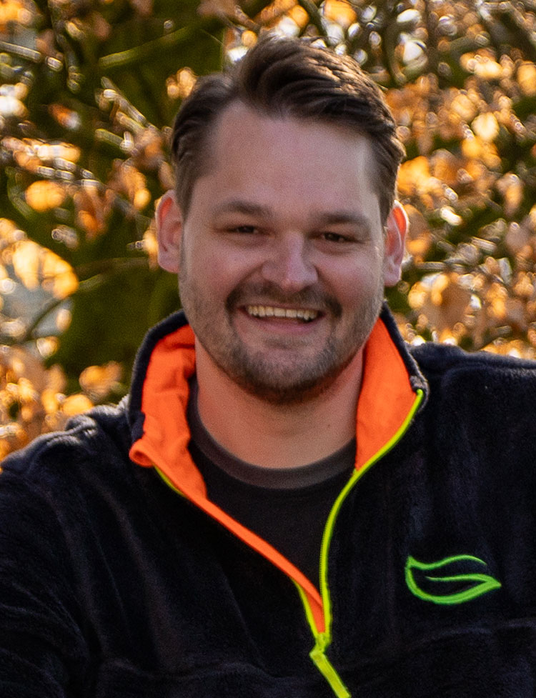 Ein Mann mit einer dunklen Jacke mit orangenen und grünen Akzenten lächelt in die Kamera.